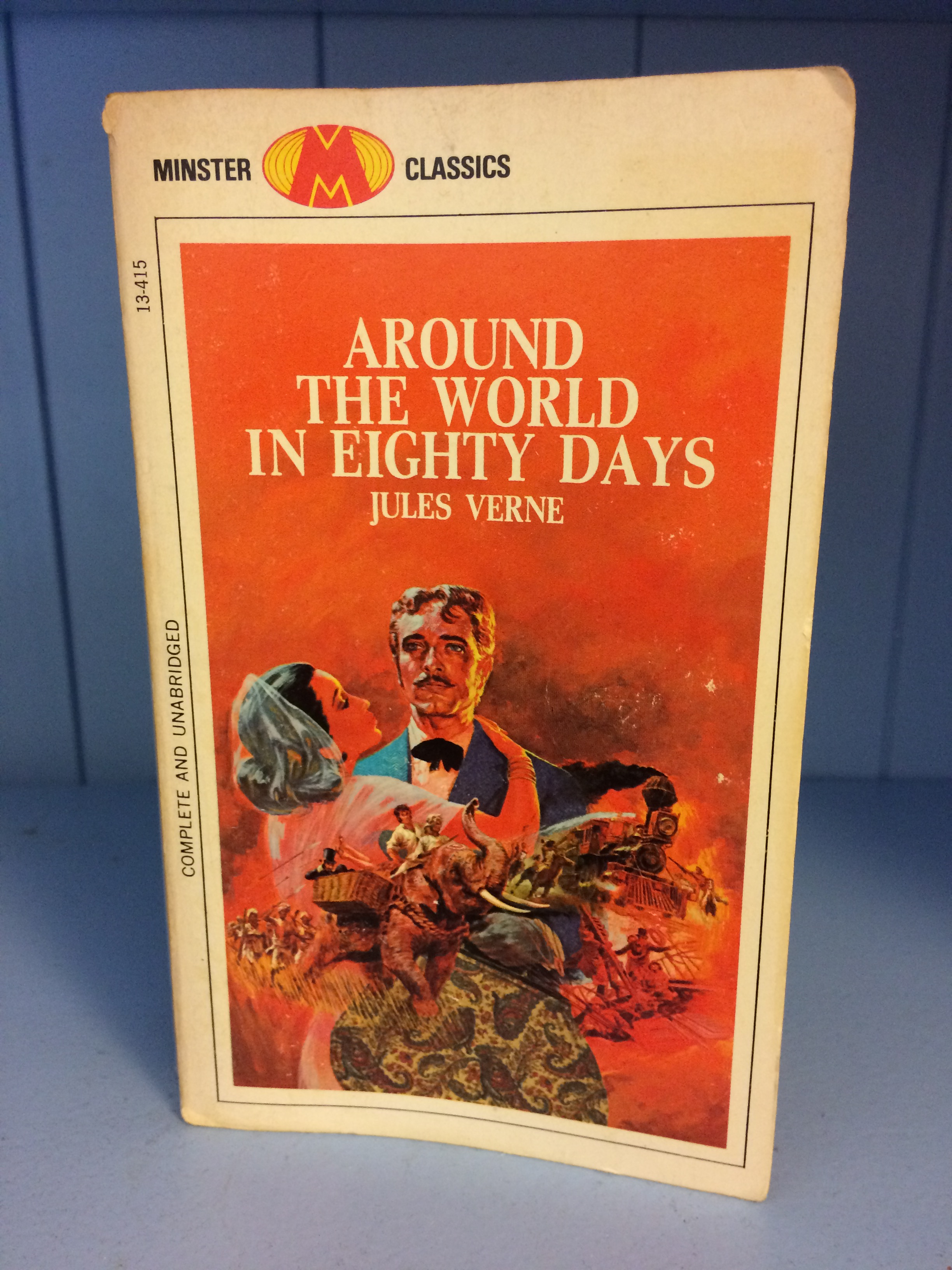 A book stood up on a blue shelf. The cover is predominately orange. Around the World in Eighty Days and Jules Verne are centred in the top third of the cover. Below you can see a man in a blue suit holding a woman in a pink dress with a white, but somewhat translucent headscarf. Circling around the two you can see a group of warriors, referred to as Sioux bandits in the text, the group on an elephant, a ship, and a steam train.