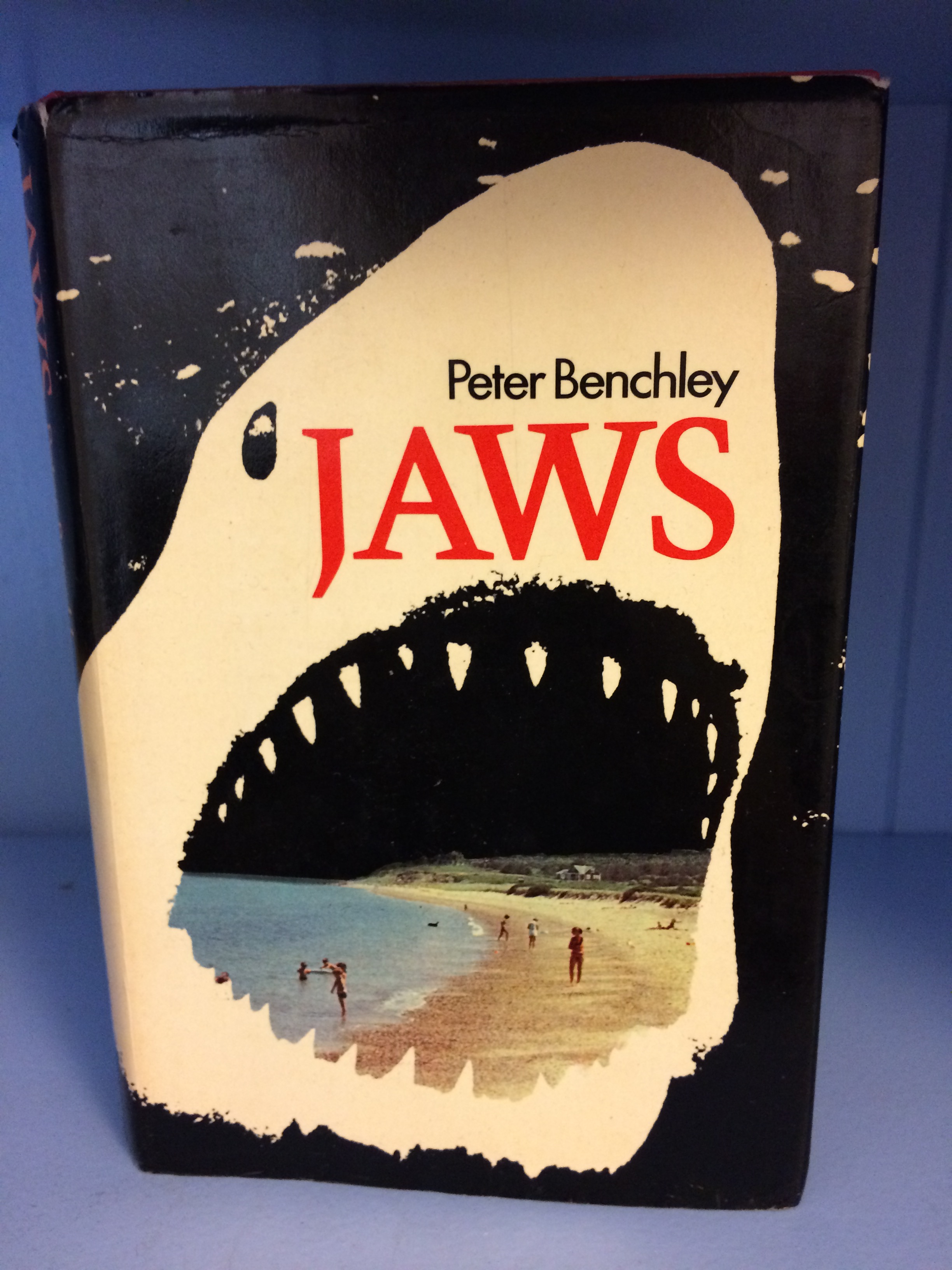 A book stood up on a blue shelf. The cover is mostly black and white with the white being the Shark. Peter Benchley and Jaws are in a large typeface on the Sharks face, JAWS is in a bolder red font. In the mouth of the shark is Amity beach, some children are swimming in the water, and others can be seen on the beach.