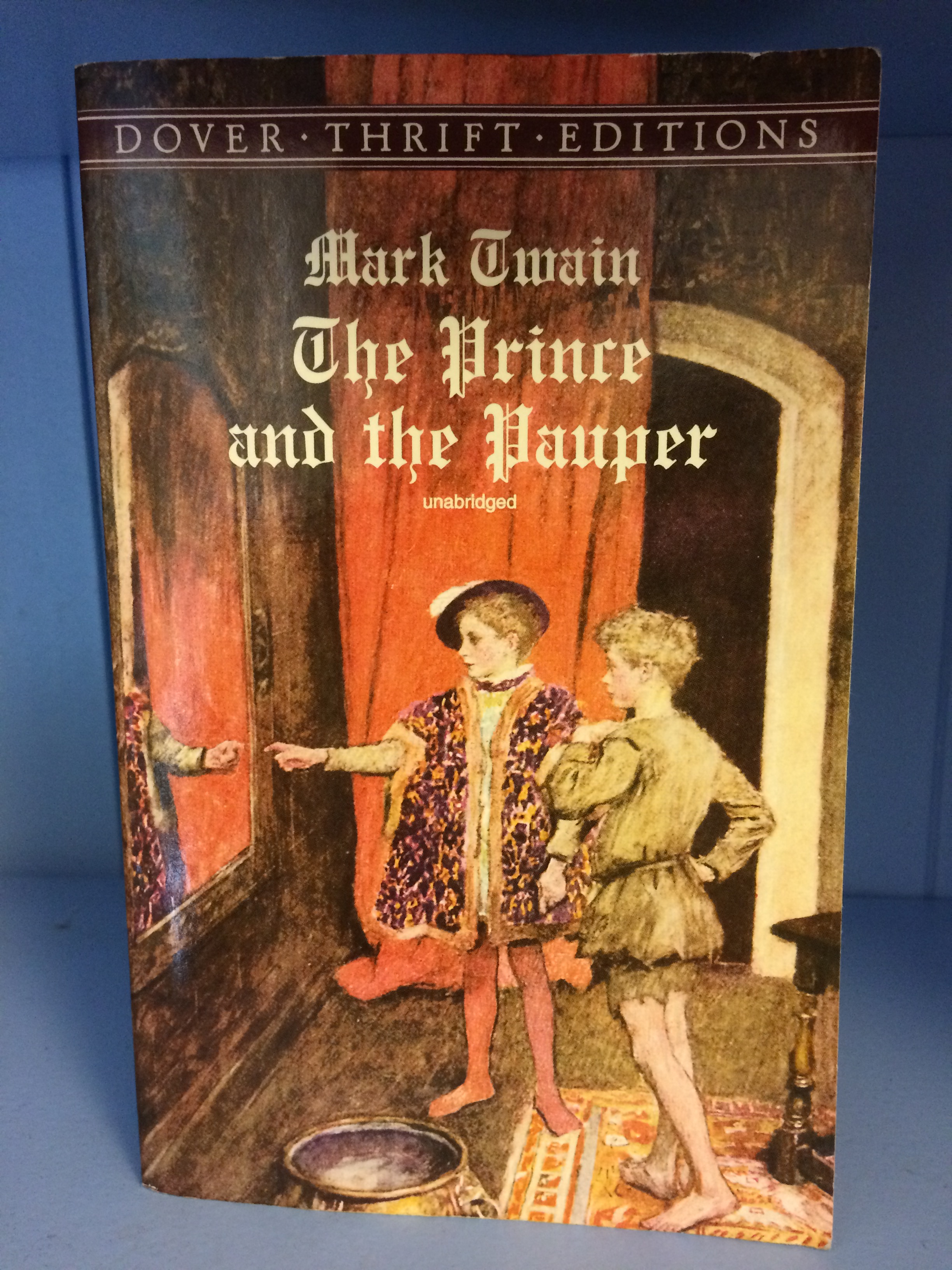 A book on a blue shelf. The text Mark Twain, The Prince and The Pauper is in a Tudor-esque font and overlaid on the image. 
The image shows two boys in front of a mirror comparing themselves. They are in the Prince's chamber. One is dressed like royalty, and the other like a pauper with bare legs.